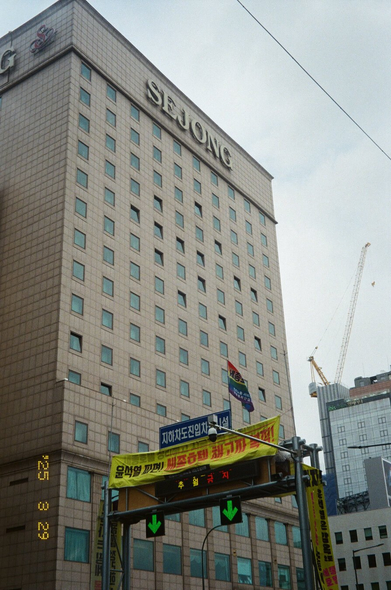 서울 세종호텔 앞 도로표지판에서 고공농성 중인 고진수 동지. 전광판 위에 “윤석열 파면! 세종호텔 해고자 복직!”이라는 현수막이 걸려있고 “세종호텔지부”라고 써있는 무지개 깃발이 나부끼고 있다.