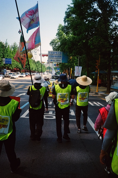 인천광역시청, 인천광역시의회 방향을 알리는 도로표지판 아래로 형광노란색 조끼와 몸자보를 입은 행진 대오가 행진하고 있다.