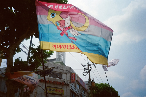 휘날리고 있는 "퀴어마법소녀연대" 깃발. 퀴어를 상징하는 배경에 각종 마법봉이 그려져있다. "만국의 마법소녀여 단결하라!"
