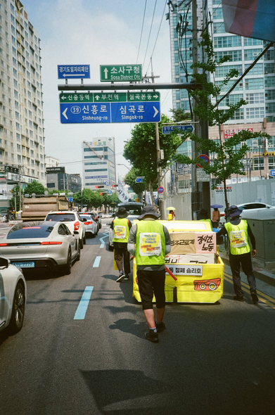 부천시의 도로표지판 아래를 행진하는 사람들과 택시 모형. "소사구청", "신흥로", "심곡로", "부천역" 등이 써있다.