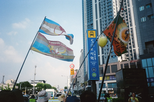 소사역 표지 앞에서 휘날리는 "퀴어마법소녀연대" 깃발과 "황금 연어알줍기 아르바이트 노동조합" 깃발.